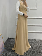 A woman wearing a beige sleeveless maxi dress over a white long-sleeve top, standing in an indoor room with a gray carpet and white wall molding.