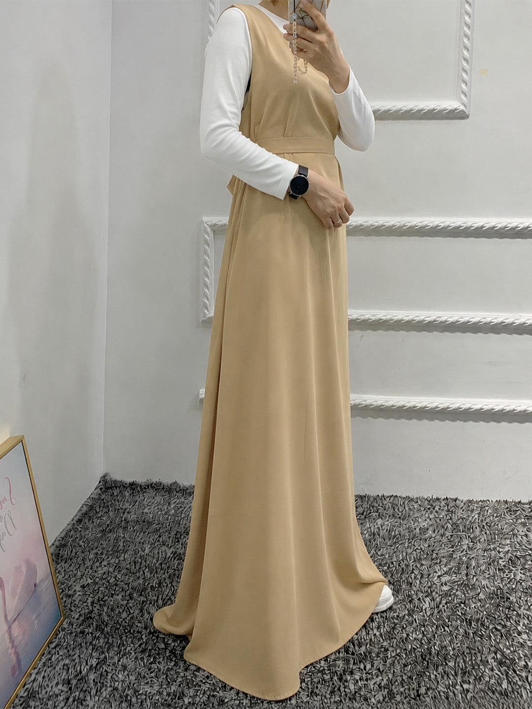 A woman wearing a beige sleeveless maxi dress over a white long-sleeve top, standing in an indoor room with a gray carpet and white wall molding.
