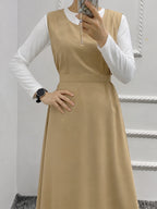 A woman wearing a beige sleeveless midi dress over a white long-sleeve collared shirt, holding a phone to take a mirror selfie in an indoor setting.