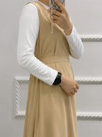 A person wearing a beige sleeveless pinafore-style dress over a white long-sleeve top, holding a phone in front of their face. The background is a white paneled wall.