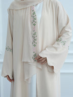 A cream-colored traditional outer robe with delicate green and pink floral embroidery, worn over a white inner garment. The image is cropped to show the upper body and hands of a model wearing the garment.