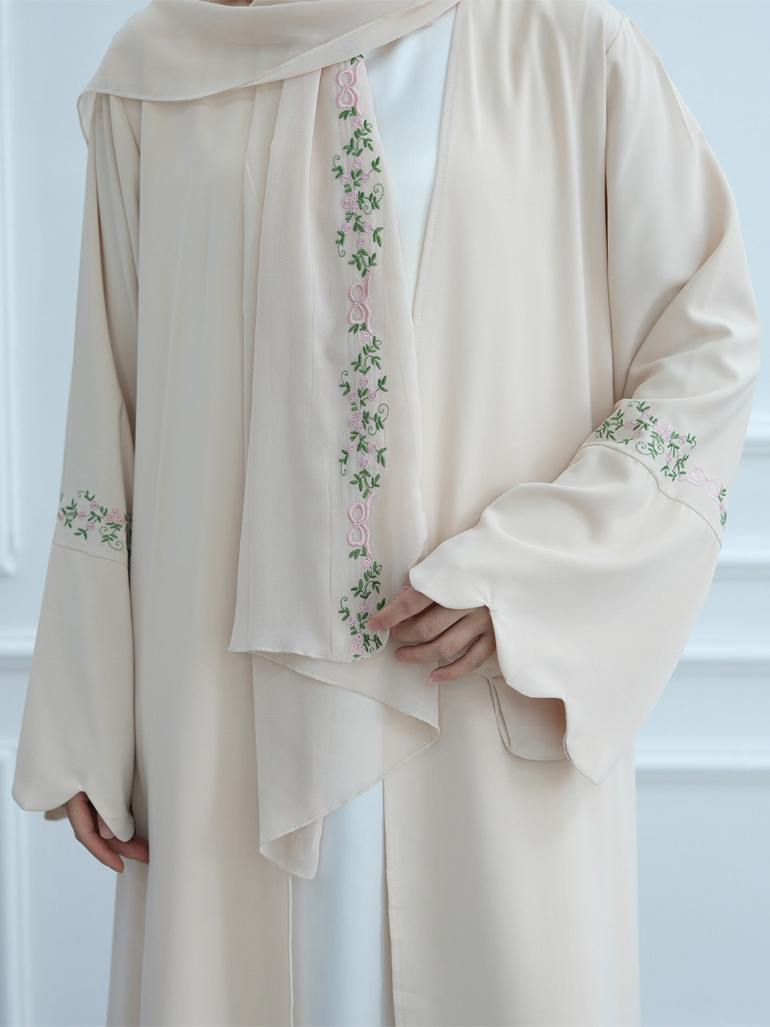 A cream-colored traditional outer robe with delicate green and pink floral embroidery, worn over a white inner garment. The image is cropped to show the upper body and hands of a model wearing the garment.