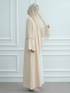 A woman wearing a modest cream-colored abaya and matching hijab with delicate green floral embroidery, standing against a white paneled wall.