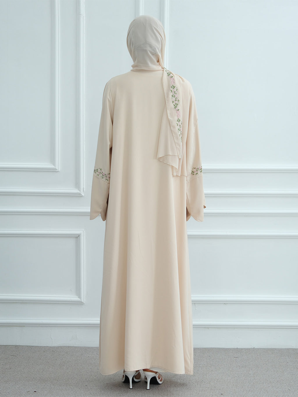 A woman viewed from behind wearing a long beige abaya dress with a matching hijab, featuring delicate floral embroidery on the back and sleeves, set against a white paneled wall.