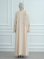 A woman viewed from behind wearing a long beige abaya dress with a matching hijab, featuring delicate floral embroidery on the back and sleeves, set against a white paneled wall.