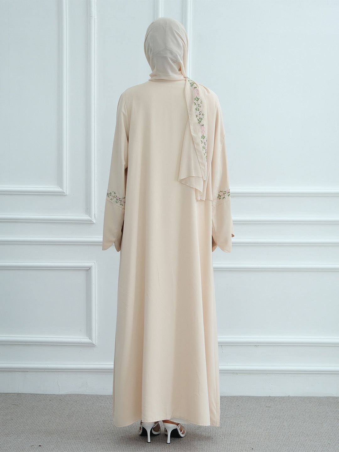 A woman viewed from behind wearing a long beige abaya dress with a matching hijab, featuring delicate floral embroidery on the back and sleeves, set against a white paneled wall.