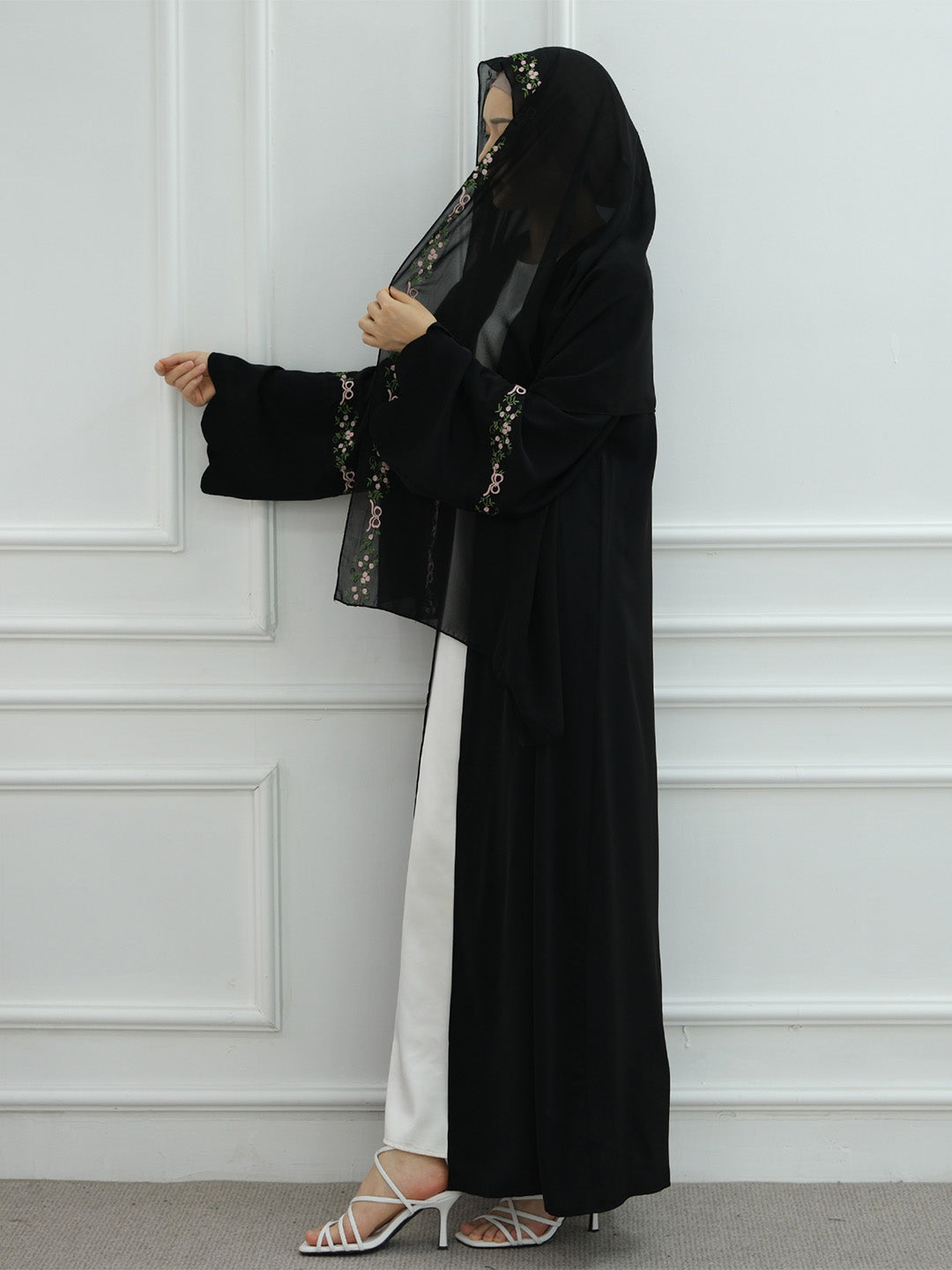 A woman modeling a black modest outfit consisting of a long abaya, a matching hijab with embroidered details, and white trousers, standing against a white paneled wall.