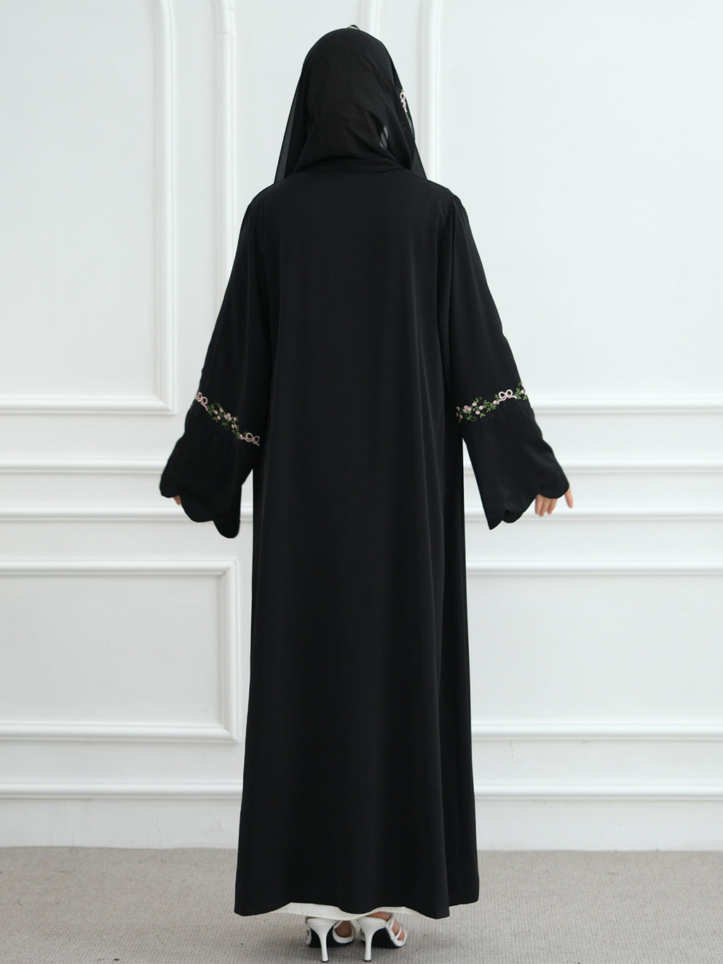 A woman viewed from behind wearing a long black abaya with a matching black head covering. The sleeves have decorative gold and white patterned trim near the cuffs.