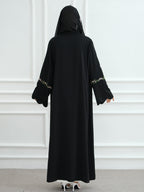 A woman viewed from behind wearing a long black abaya with a matching black head covering. The sleeves have decorative gold and white patterned trim near the cuffs.