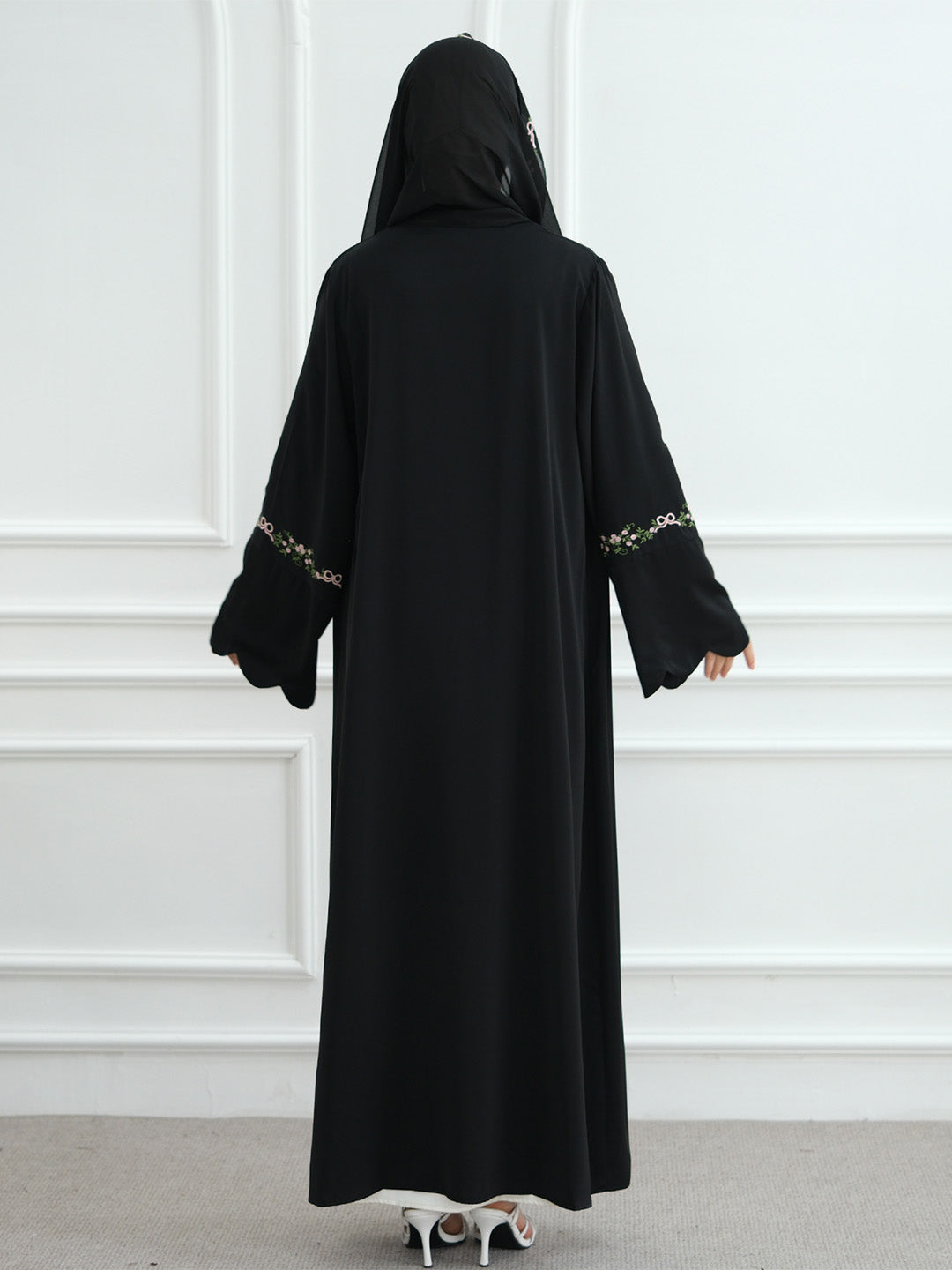 A woman viewed from behind wearing a long black abaya with a matching black head covering. The sleeves have decorative gold and white patterned trim near the cuffs.
