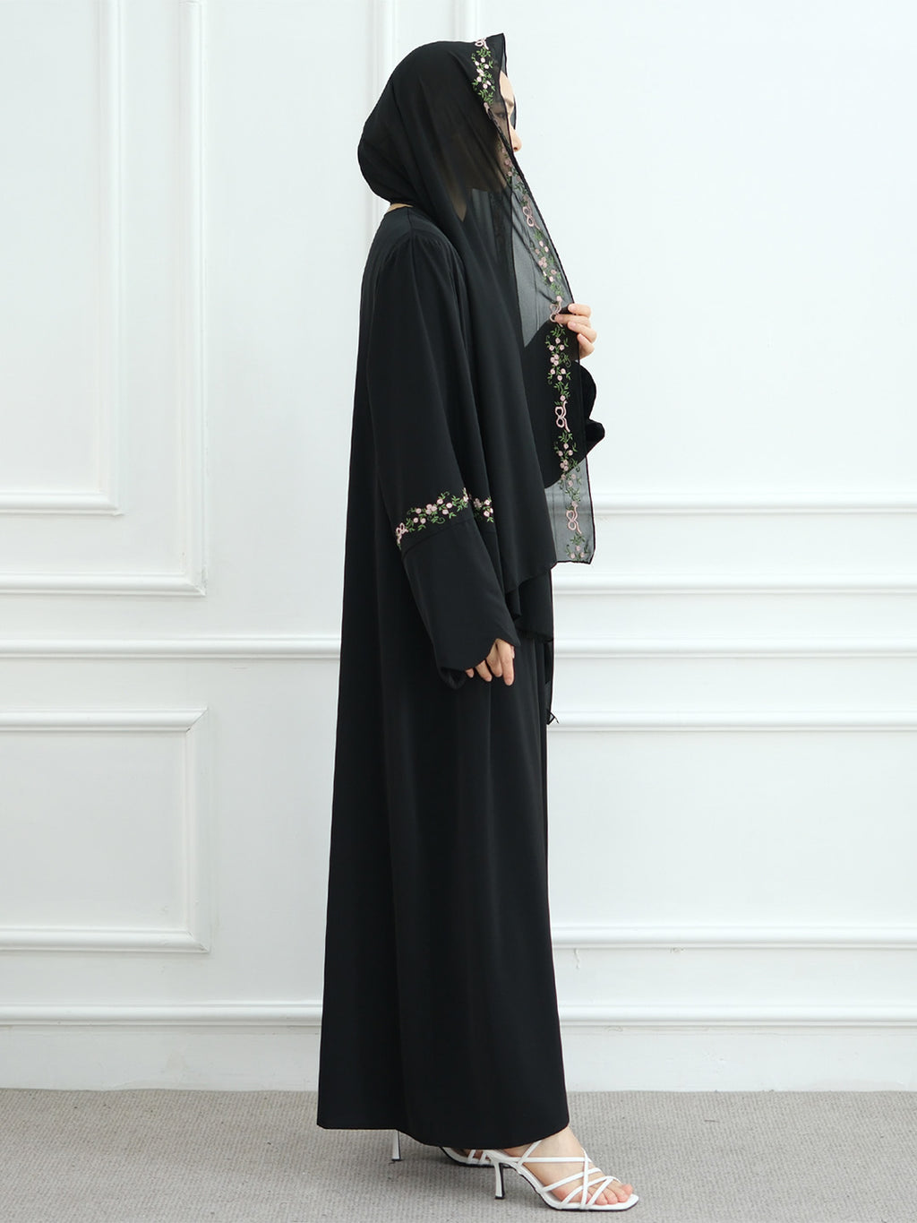 A woman modeling a black modest outfit consisting of a long abaya with embroidered details on the sleeves, a matching hijab, and white high-heeled sandals, standing against a white paneled wall.
