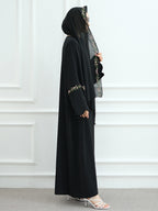 A woman modeling a black modest outfit consisting of a long abaya with embroidered details on the sleeves, a matching hijab, and white high-heeled sandals, standing against a white paneled wall.