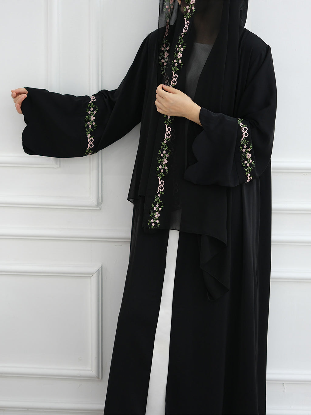 A woman wearing a black abaya with ornate floral embroidery in green, white, and gold along the front opening and sleeve cuffs, paired with a black head covering, standing against a white paneled wall.