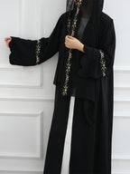 A woman wearing a black abaya with ornate floral embroidery in green, white, and gold along the front opening and sleeve cuffs, paired with a black head covering, standing against a white paneled wall.