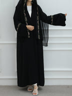 A woman wearing a long black abaya with ornate gold embroidery on the trim and cuffs, a matching black headscarf, a white inner top, and white strappy sandals, standing against a white paneled wall.