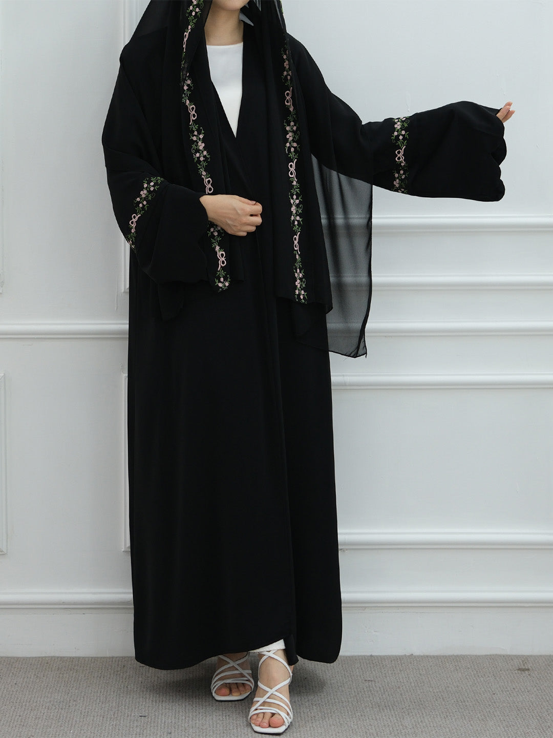 A woman wearing a long black abaya with ornate gold embroidery on the trim and cuffs, a matching black headscarf, a white inner top, and white strappy sandals, standing against a white paneled wall.