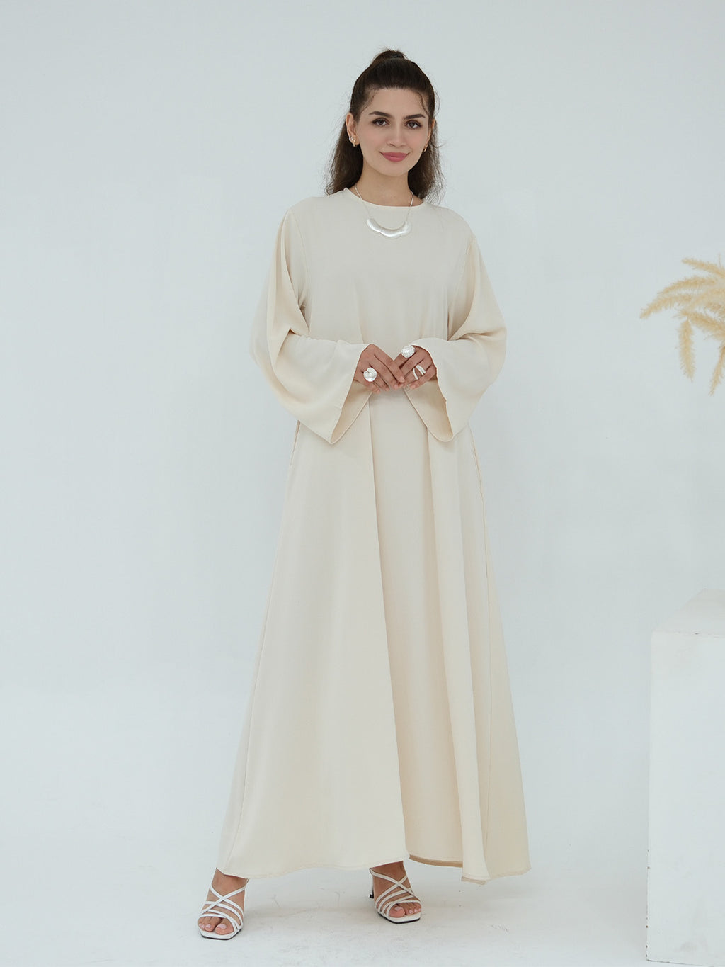A woman modeling a long, flowing abaya dress in a light cream or beige color, featuring wide bell sleeves and a round neckline, paired with white strappy high-heeled sandals against a clean white studio background.