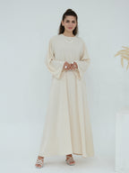 A woman modeling a long, flowing abaya dress in a light cream or beige color, featuring wide bell sleeves and a round neckline, paired with white strappy high-heeled sandals against a clean white studio background.
