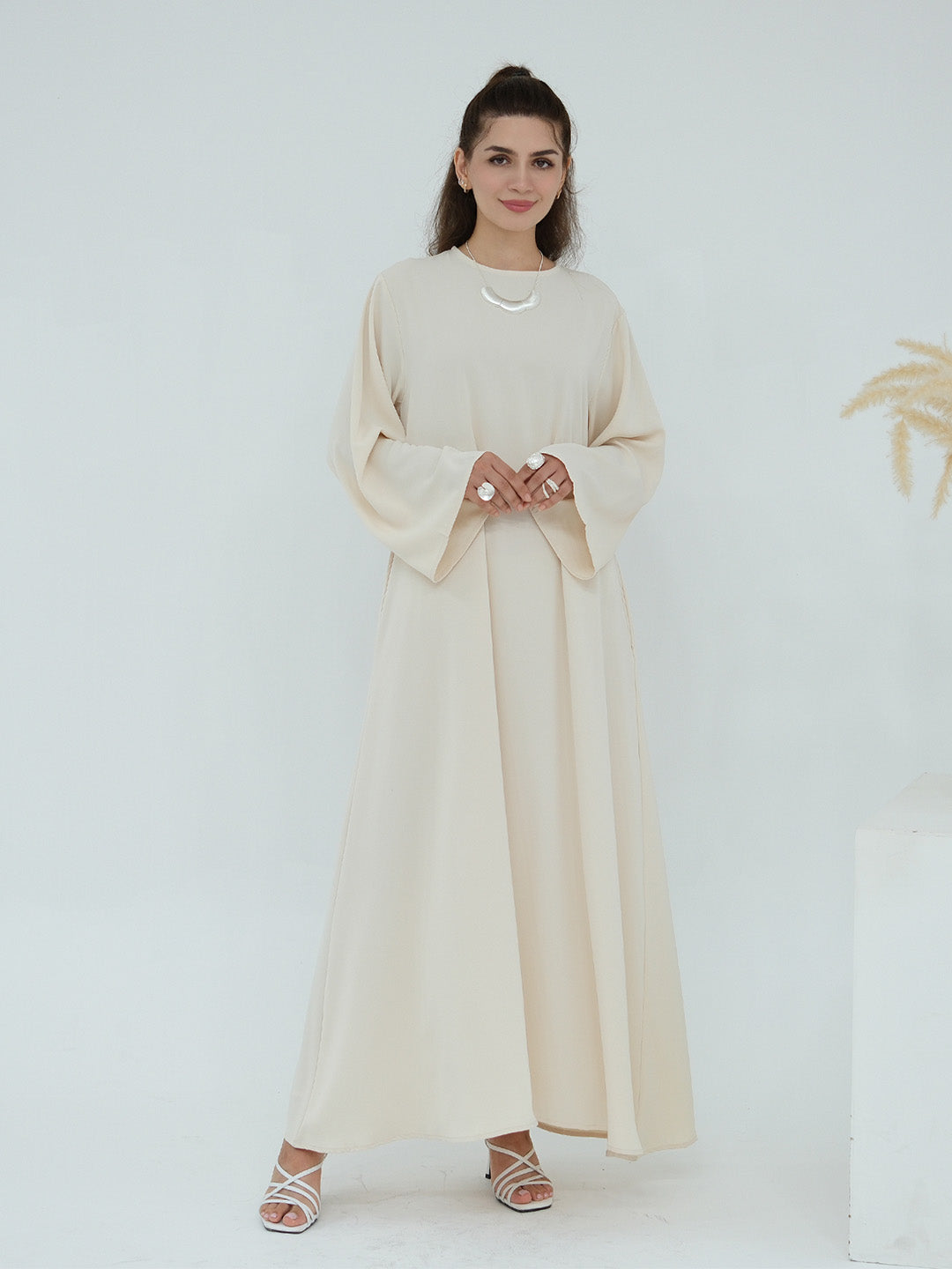 A woman modeling a long, flowing abaya dress in a light cream or beige color, featuring wide bell sleeves and a round neckline, paired with white strappy high-heeled sandals against a clean white studio background.
