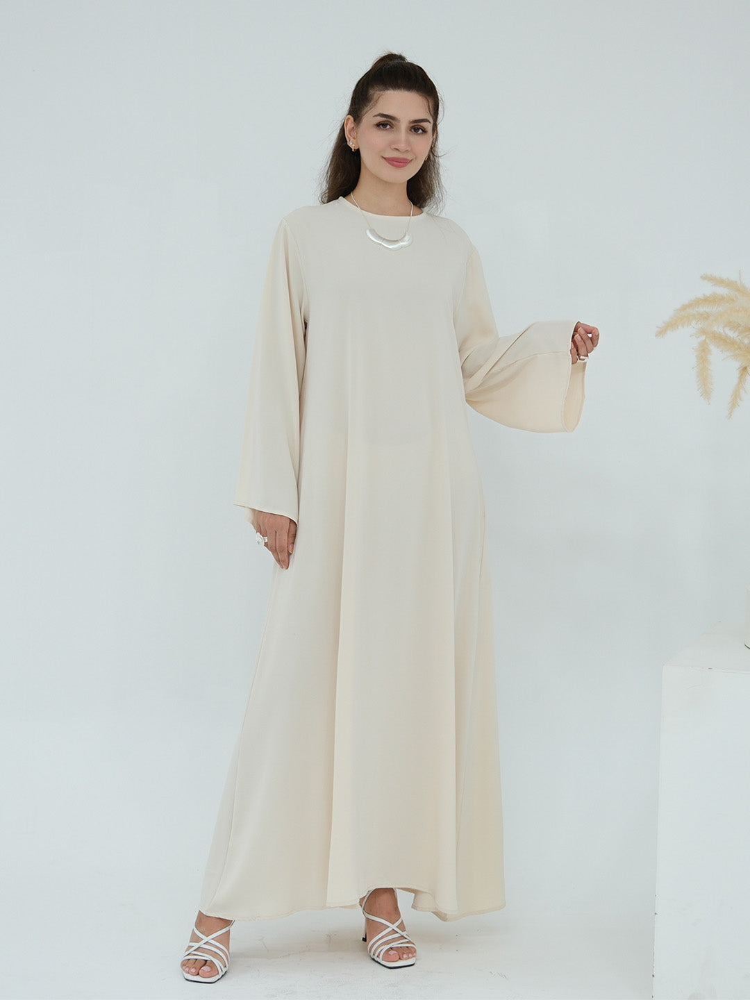 A woman modeling a long-sleeved, floor-length beige dress with a round neckline and a small silver necklace, standing against a clean white studio background.