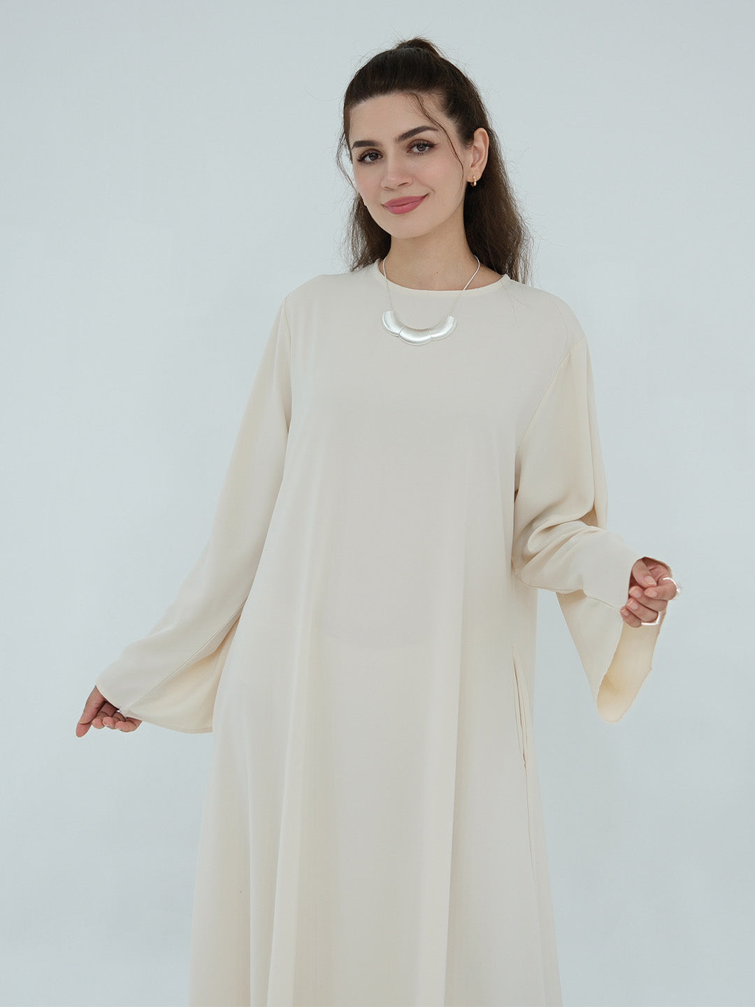 A woman modeling a long-sleeved, loose-fitting cream-colored dress or kaftan against a plain light gray studio background.