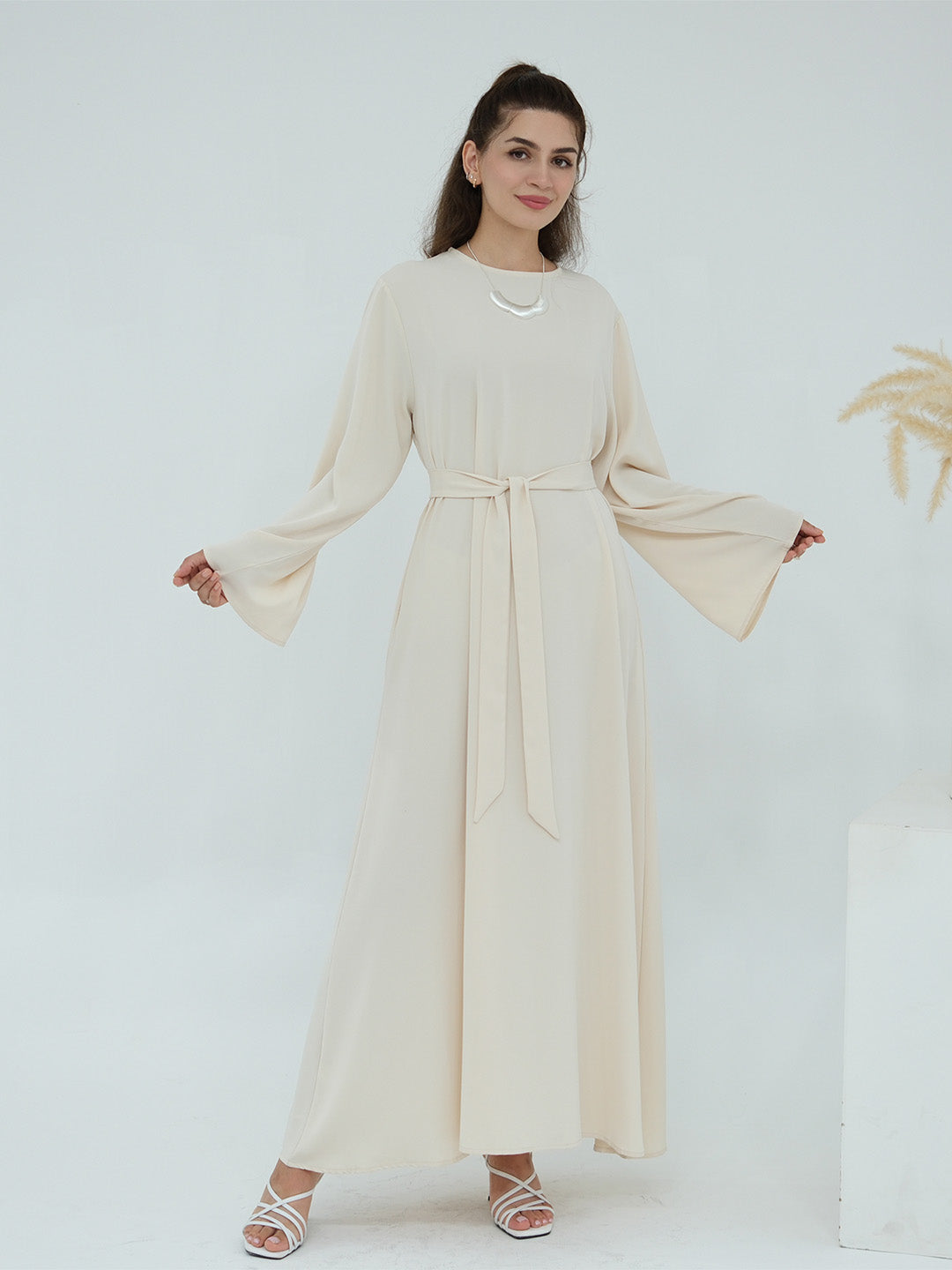 A woman modeling a long-sleeved cream-colored maxi dress with a matching waist tie belt, set against a clean white studio background.