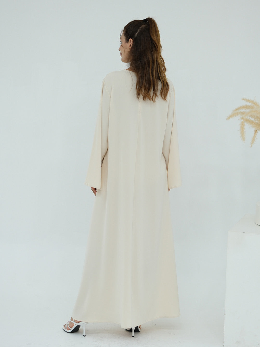 A woman viewed from behind wearing a long, flowing cream-colored abaya dress with wide sleeves, paired with silver high-heeled sandals, set against a clean white studio background.