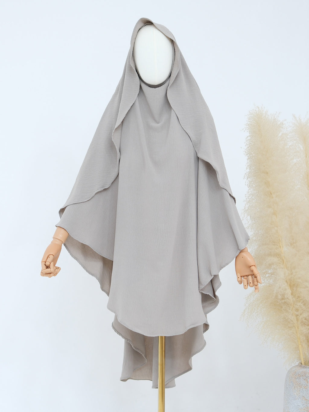 A light gray modest garment with a draped hood and wide flowing sleeves, displayed on a mannequin against a clean white background with decorative pampas grass on the right.
