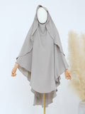 A light gray modest garment with a draped hood and wide flowing sleeves, displayed on a mannequin against a clean white background with decorative pampas grass on the right.