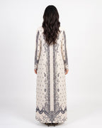 A woman viewed from the back wearing a long cream-colored kaftan dress with dark gray and black floral embroidery and vertical border details, set against a plain white studio background.