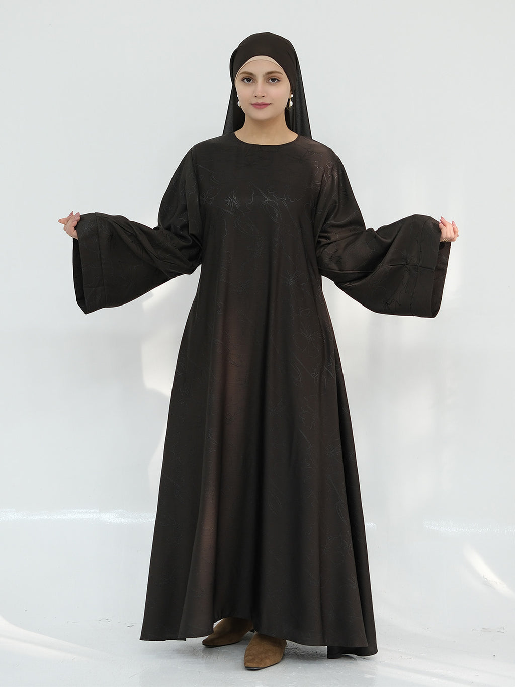 A woman modeling a long, loose-fitting black abaya dress with wide bell sleeves and a matching black headscarf, standing against a plain white studio background.