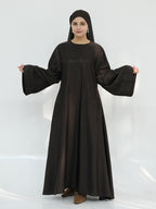 A woman modeling a long, loose-fitting black abaya dress with wide bell sleeves and a matching black headscarf, standing against a plain white studio background.