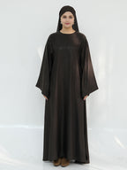 A woman wearing a long, loose-fitting dark brown abaya dress with wide sleeves and a matching head covering, standing against a plain white studio background.