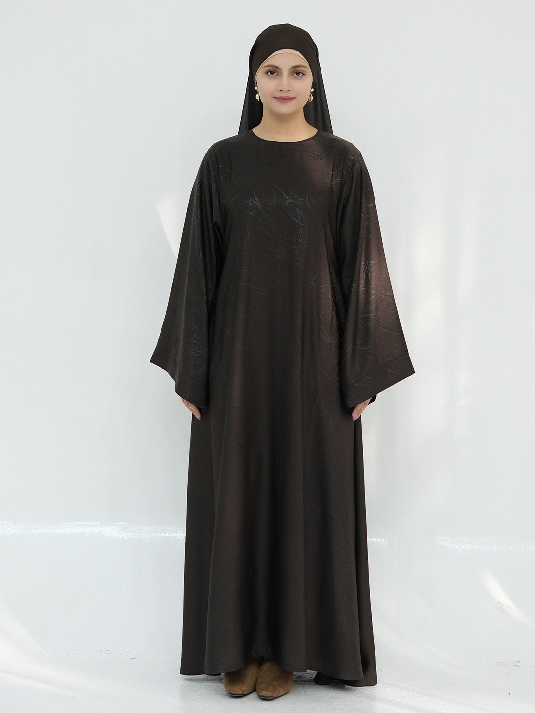 A woman wearing a long, loose-fitting dark brown abaya dress with wide sleeves and a matching head covering, standing against a plain white studio background.