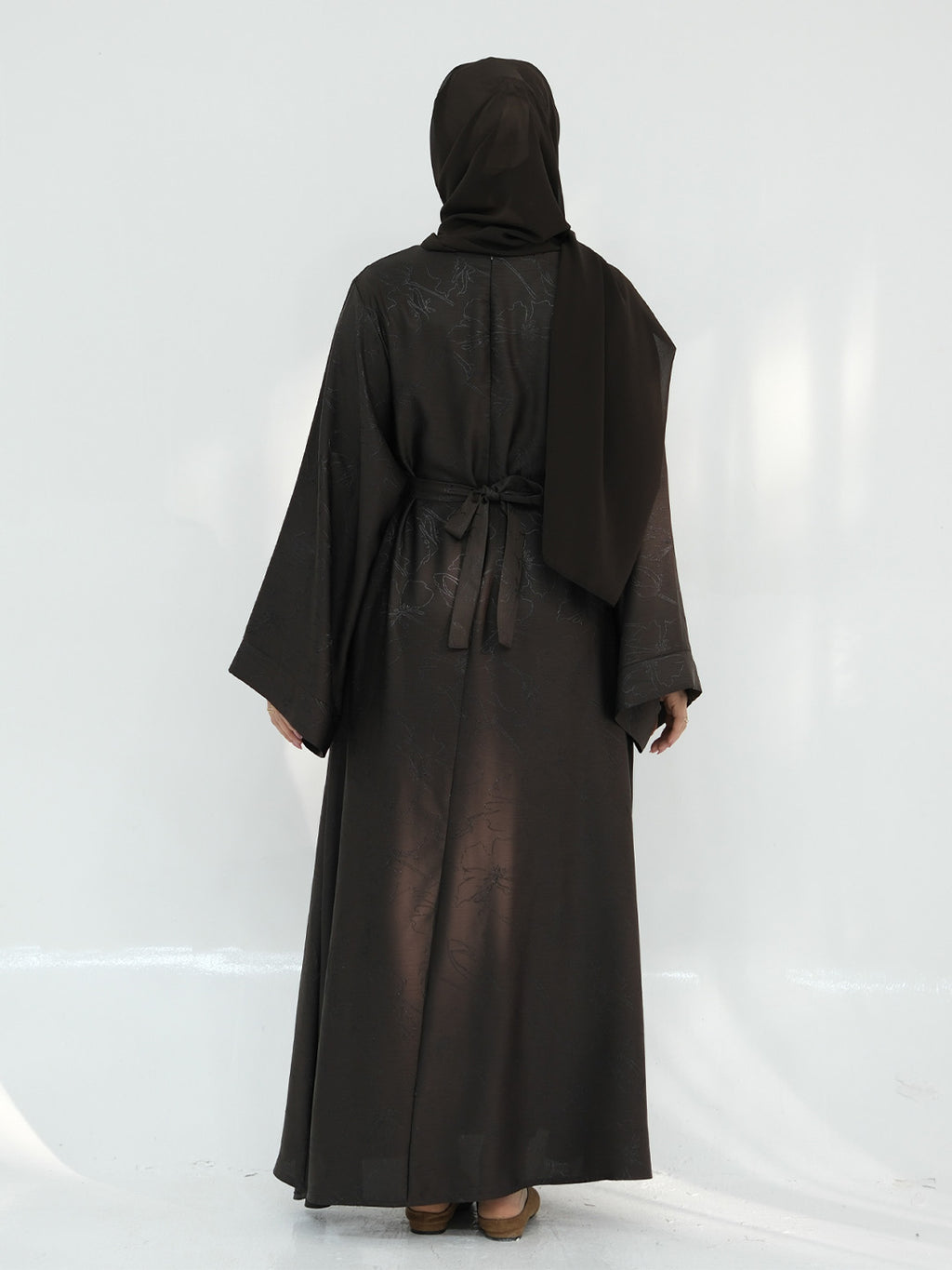 A woman wearing a long black abaya robe with a matching headscarf, shown from the back against a white studio backdrop.