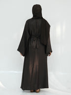A woman wearing a long black abaya robe with a matching headscarf, shown from the back against a white studio backdrop.