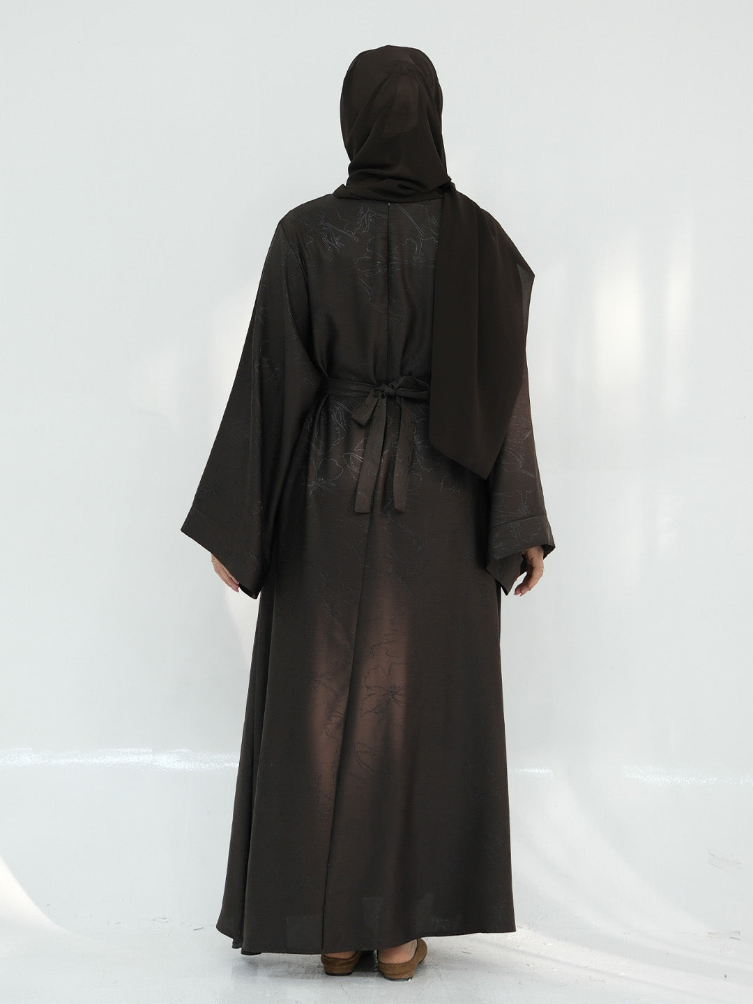 A woman wearing a long black abaya robe with a matching headscarf, shown from the back against a white studio backdrop.