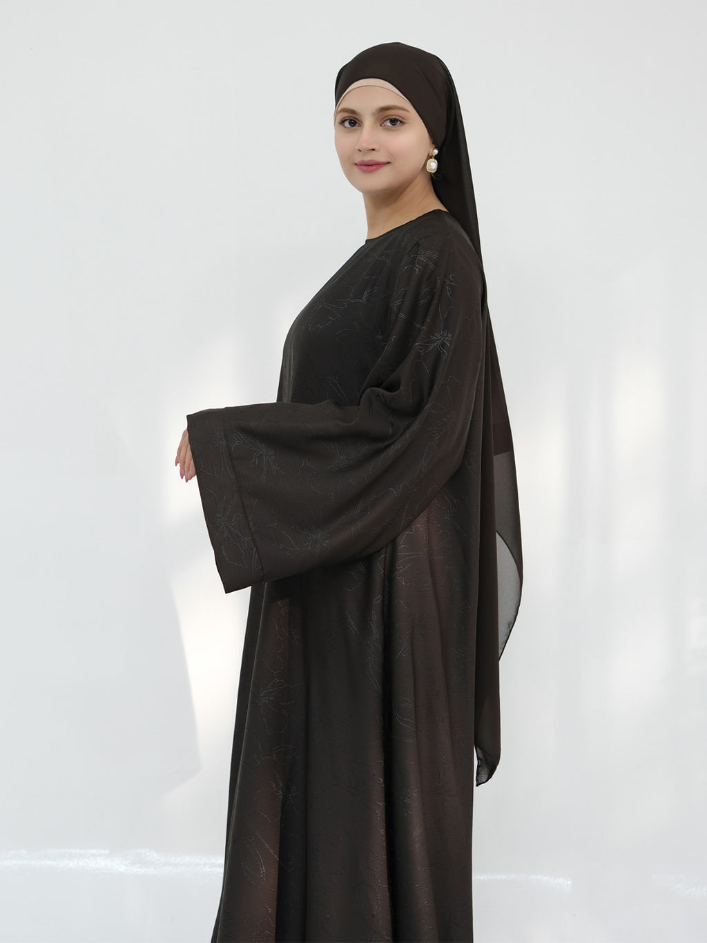 A woman modeling a dark brown abaya with matching headscarf, shown from the side against a light gray studio background.