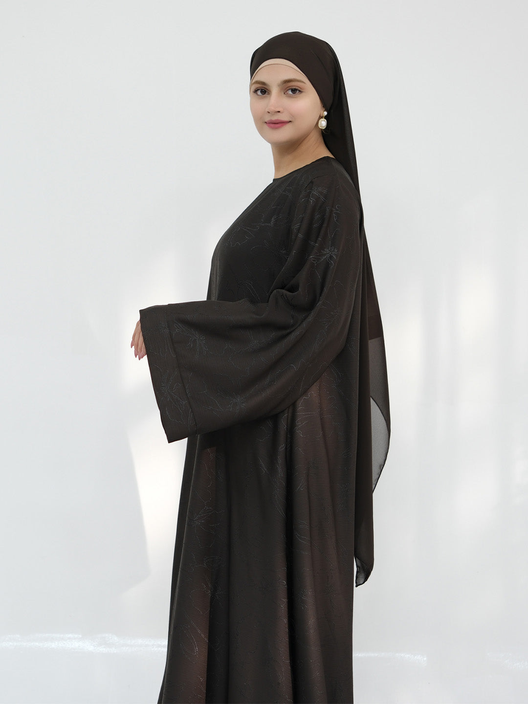A woman modeling a dark brown abaya with matching headscarf, shown from the side against a light gray studio background.