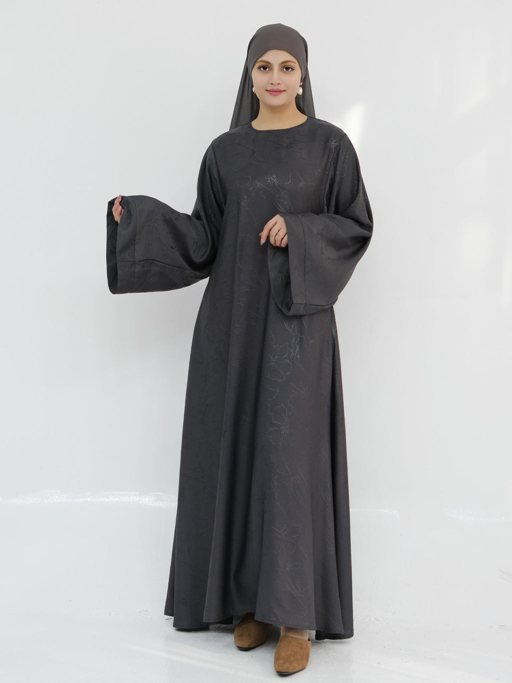 A woman modeling a long, flowing black abaya dress with wide bell sleeves and a matching black hijab, standing against a plain white studio background.