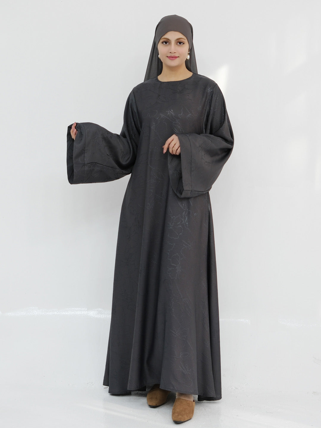 A woman modeling a long, flowing black abaya dress with wide bell sleeves and a matching black hijab, standing against a plain white studio background.