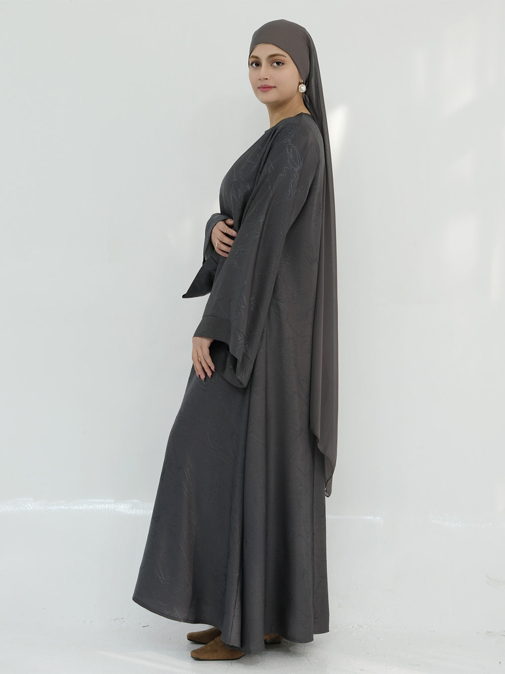 A woman modeling a dark gray modest outfit consisting of a long-sleeved abaya, a matching headscarf, and brown flat shoes, standing against a plain white studio background.