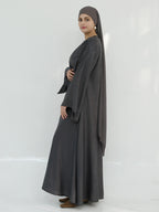 A woman modeling a dark gray modest outfit consisting of a long-sleeved abaya, a matching headscarf, and brown flat shoes, standing against a plain white studio background.