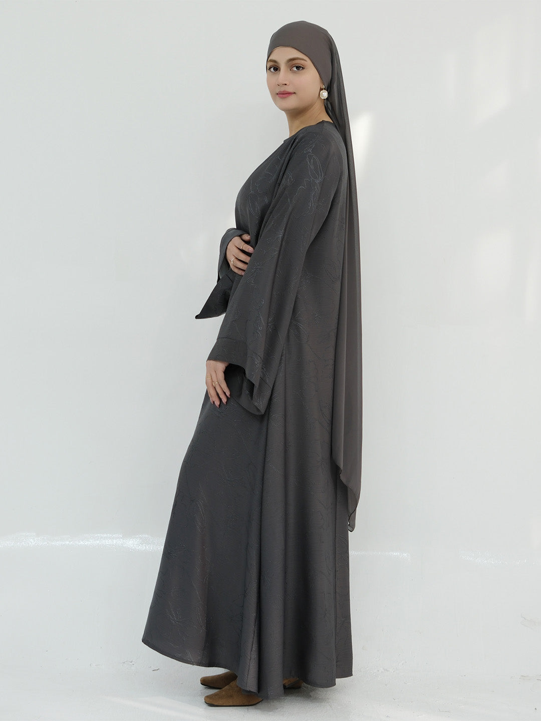 A woman modeling a dark gray modest outfit consisting of a long-sleeved abaya, a matching headscarf, and brown flat shoes, standing against a plain white studio background.