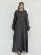 A woman wearing a long, loose-fitting dark gray abaya dress with a matching hijab, standing against a plain white studio background.