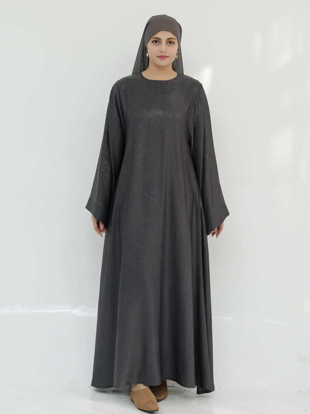A woman wearing a long, loose-fitting dark gray abaya dress with a matching hijab, standing against a plain white studio background.