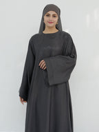 A woman wearing a dark charcoal gray abaya with a matching hijab, standing against a plain light gray background.