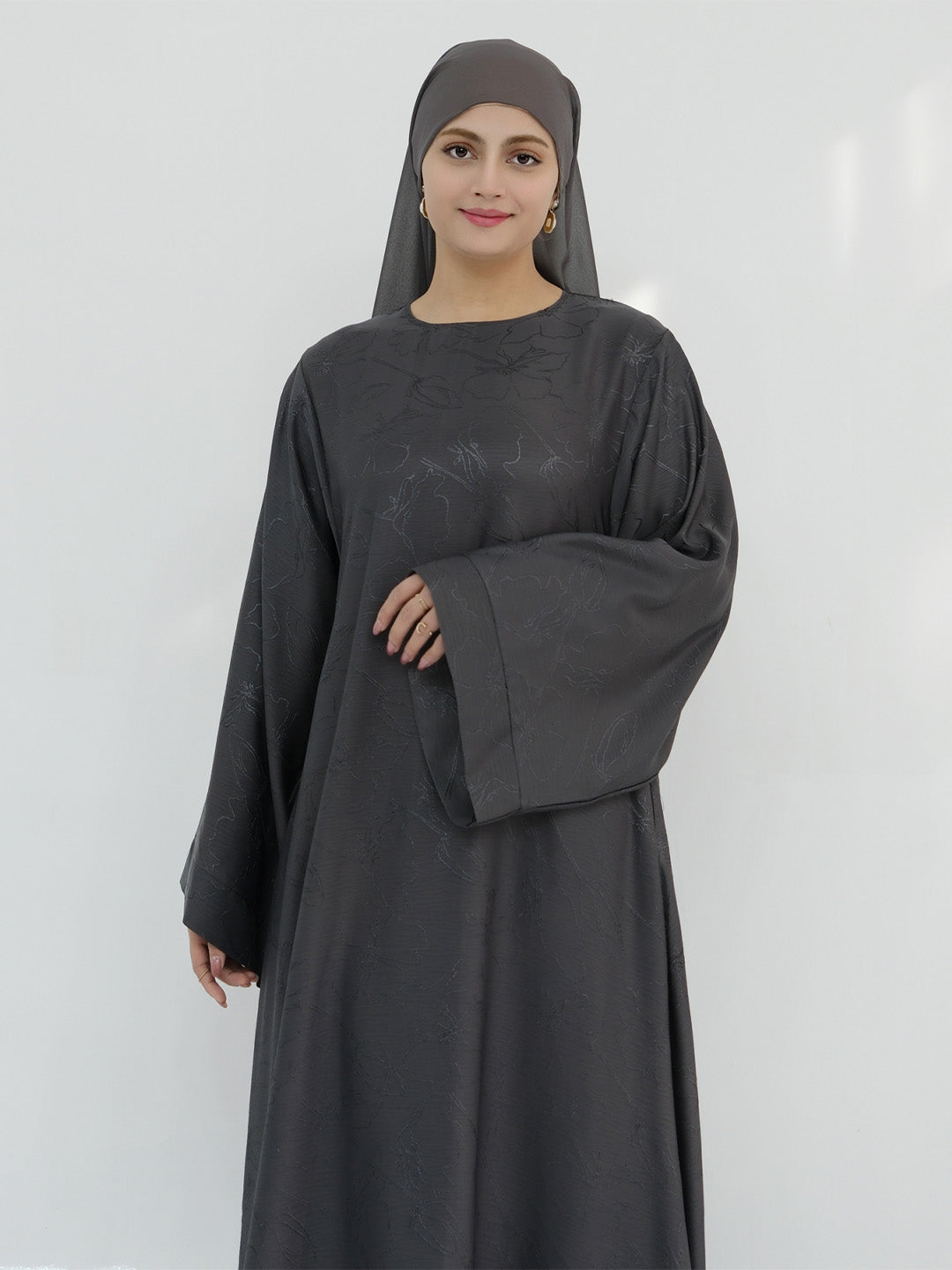 A woman wearing a dark charcoal gray abaya with a matching hijab, standing against a plain light gray background.