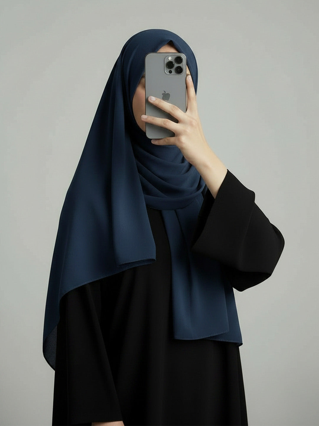 A woman wearing a dark navy blue hijab and a black dress takes a mirror selfie with her phone against a plain gray background.