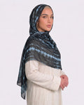 A woman wearing a modern black printed modal hijab with horizontal tie-dye stripes in steel blue and slate grey, paired with a cream outfit.